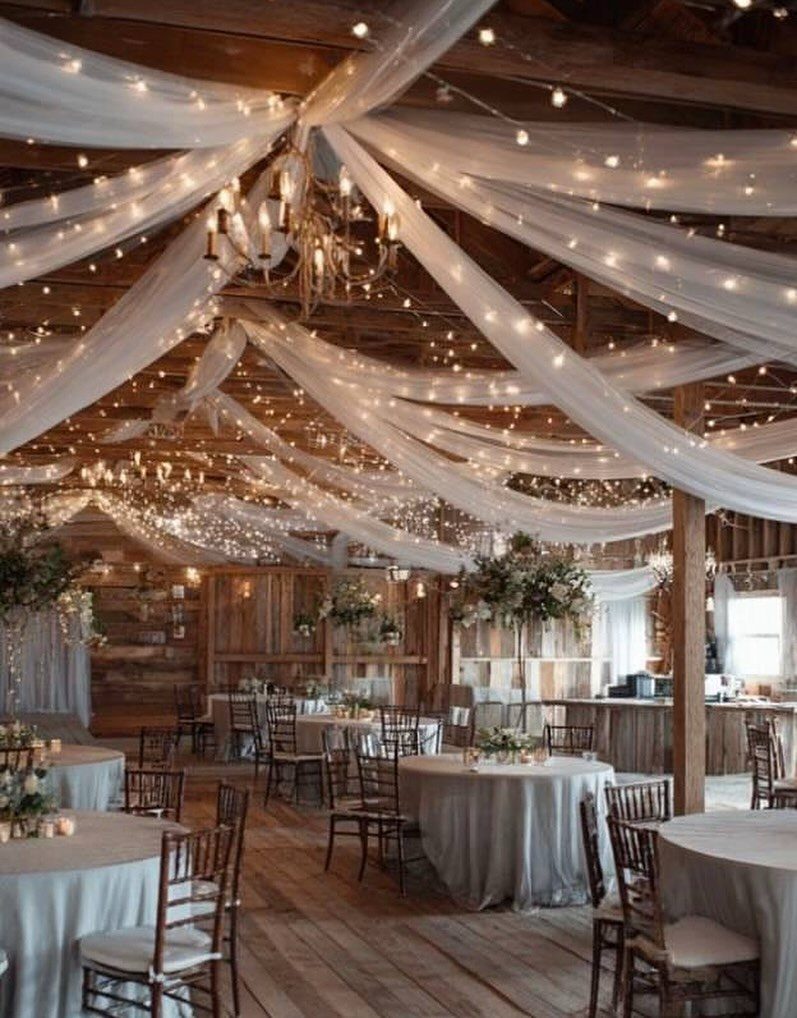 Rustic barn wedding reception with white ceiling draping, string lights, and round tables.