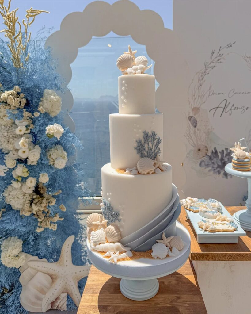 Three-tier white wedding cake decorated with seashells and coral on a blue pedestal.