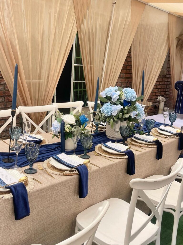 Wedding table setting with navy blue accents, beige linens, and blue floral centerpieces.