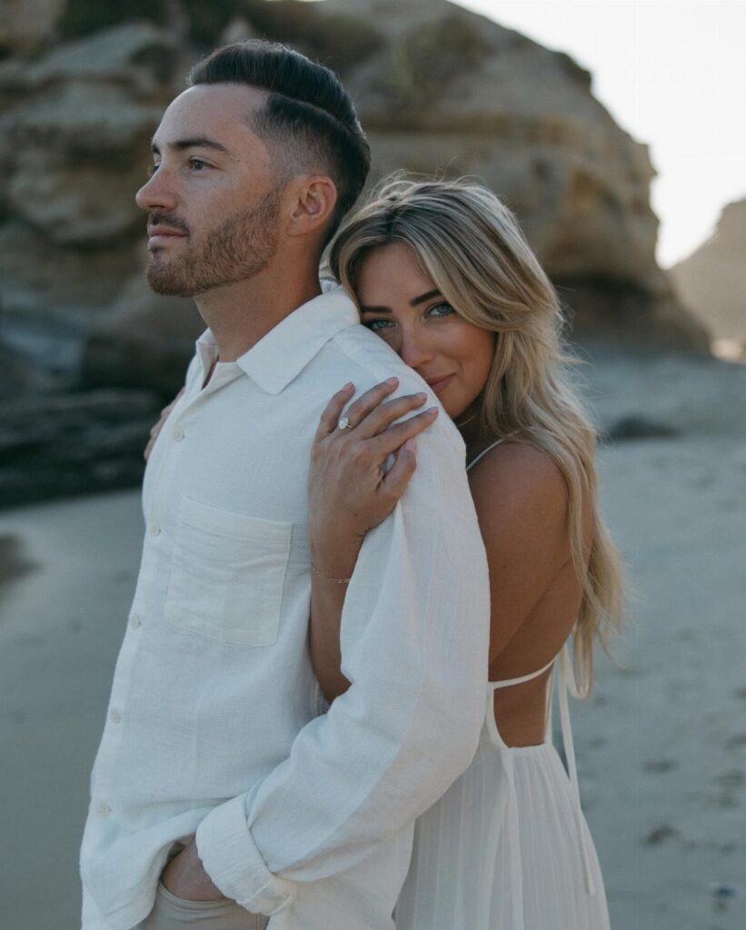 Couple in white wedding attire posing together on a sandy beach with rocks.