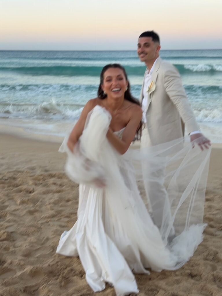 Bride and groom smiling and walking on a sandy beach by the ocean.