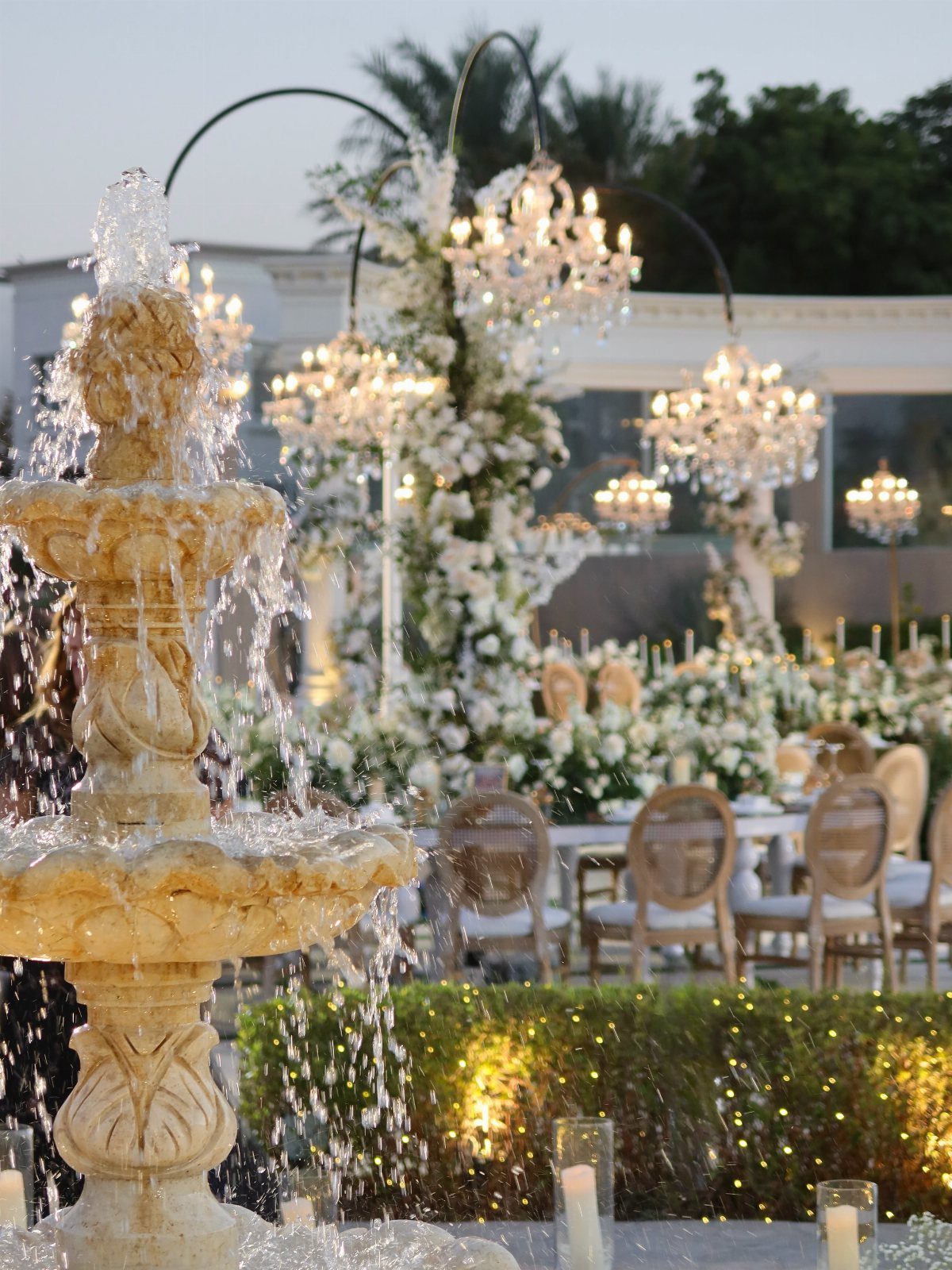 Tiered stone fountain and elegant outdoor wedding reception with chandeliers and white flowers.