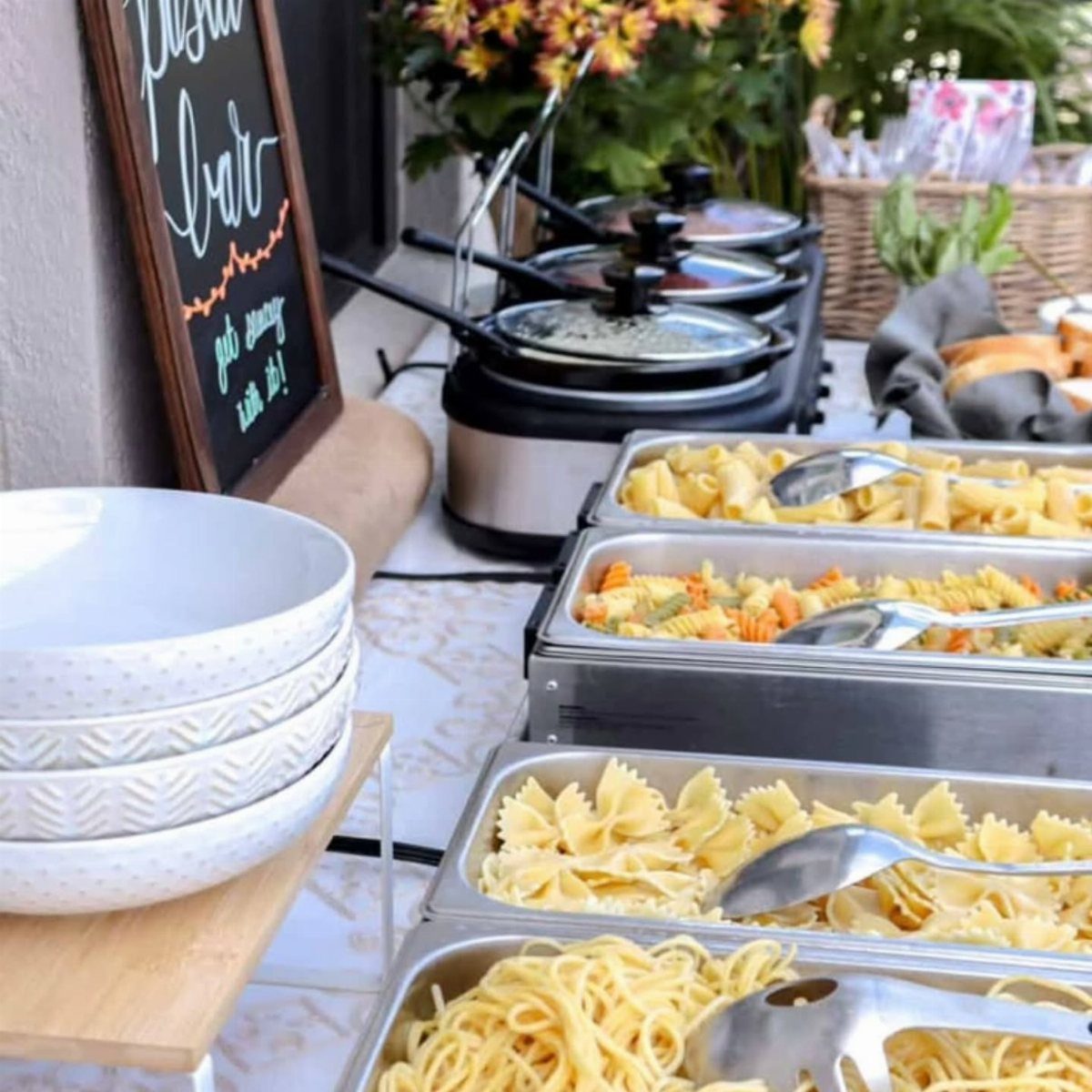 Pasta buffet with various noodle types in metal trays and warming pots.