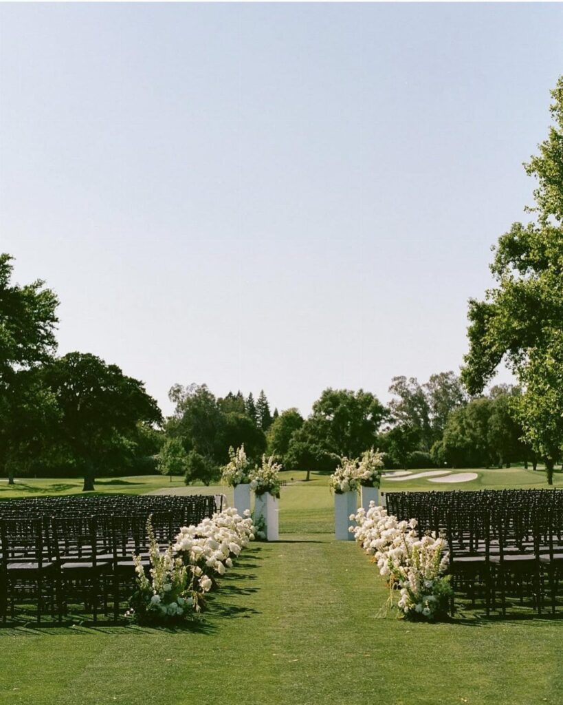 Outdoor wedding ceremony setup with dark chairs and white floral aisle arrangements on grass.