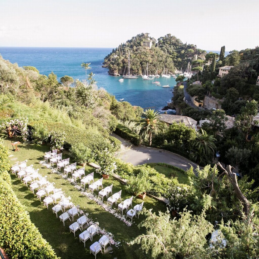 White wedding chairs arranged on a green lawn overlooking a Mediterranean coastal bay.