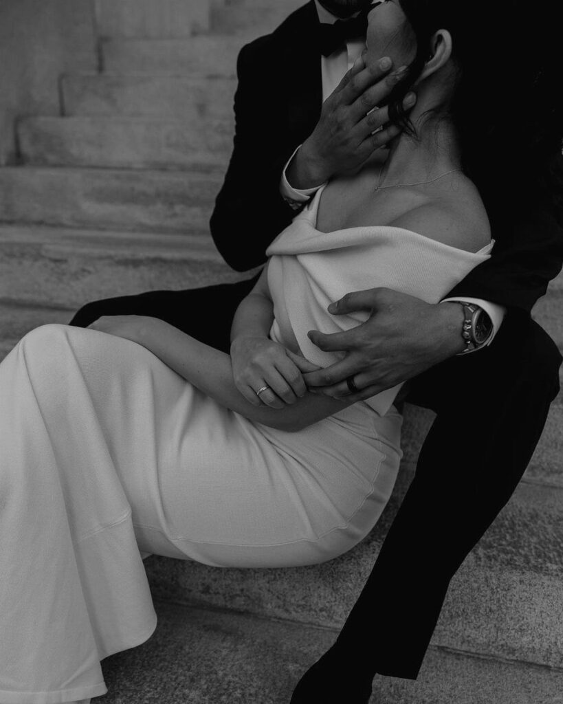 Couple in wedding attire embracing while sitting on stone steps in black and white.