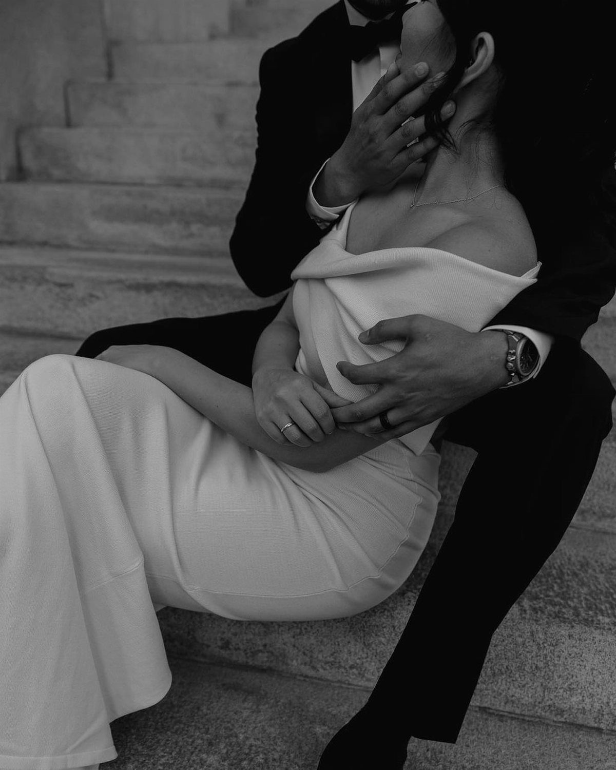 Couple in wedding attire embracing while sitting on stone steps in black and white.