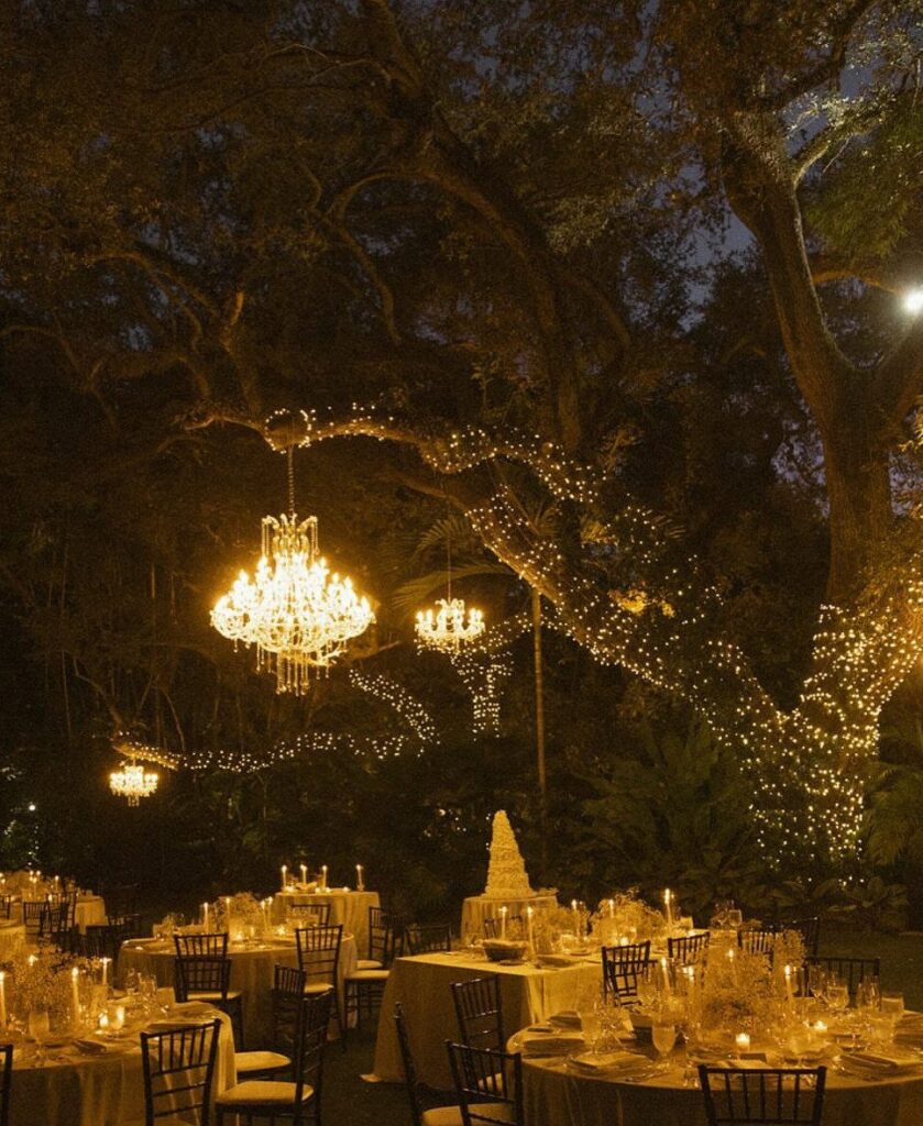 Outdoor wedding reception under a large tree with fairy lights and crystal chandeliers.