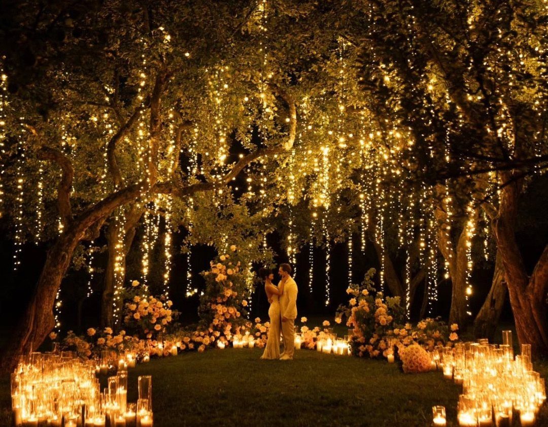 Couple standing under trees with hanging fairy lights and candles for a wedding.