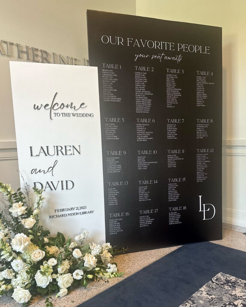 Black wedding seating chart board and white welcome sign with white floral arrangements.