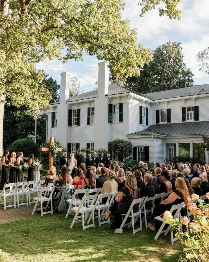 Outdoor wedding ceremony with guests in white chairs before a white colonial house.