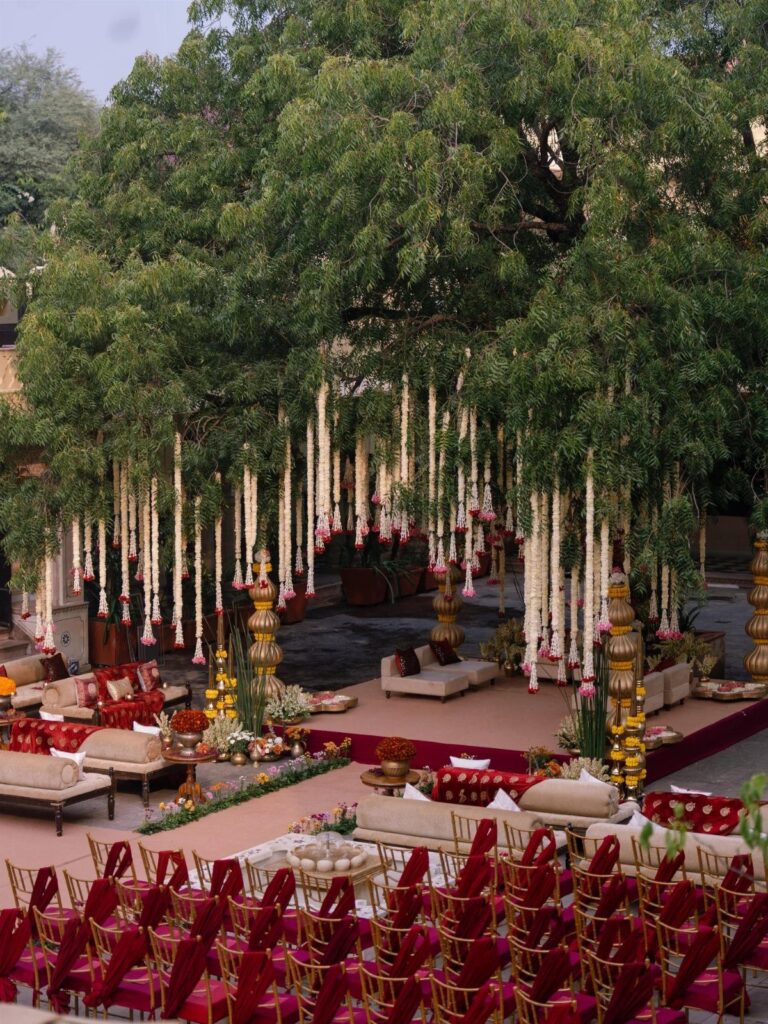 Outdoor Indian wedding venue with floral garlands hanging from a large green tree.