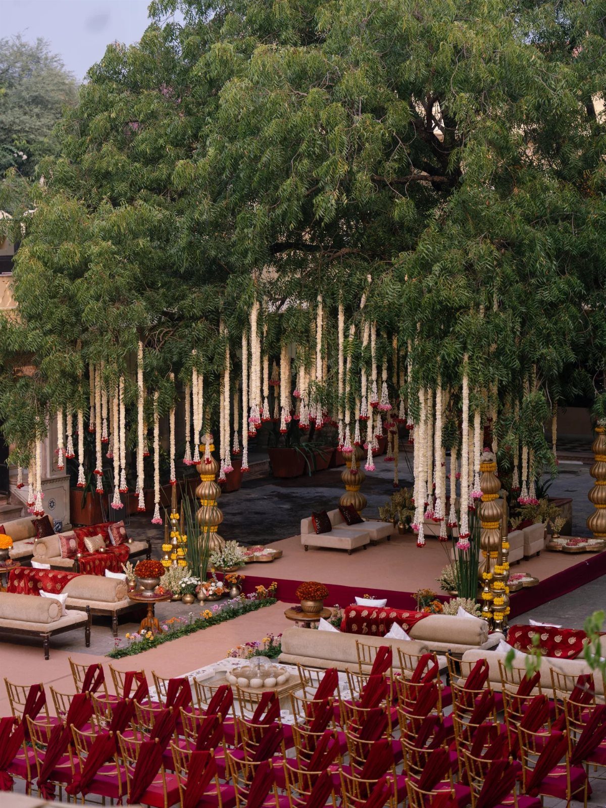 Outdoor Indian wedding venue with floral garlands hanging from a large green tree.