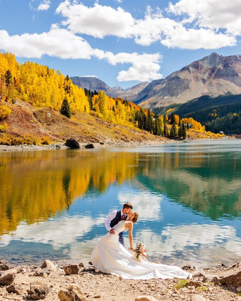 Newlywed couple kissing on a rocky shore by a mountain lake with autumn foliage.