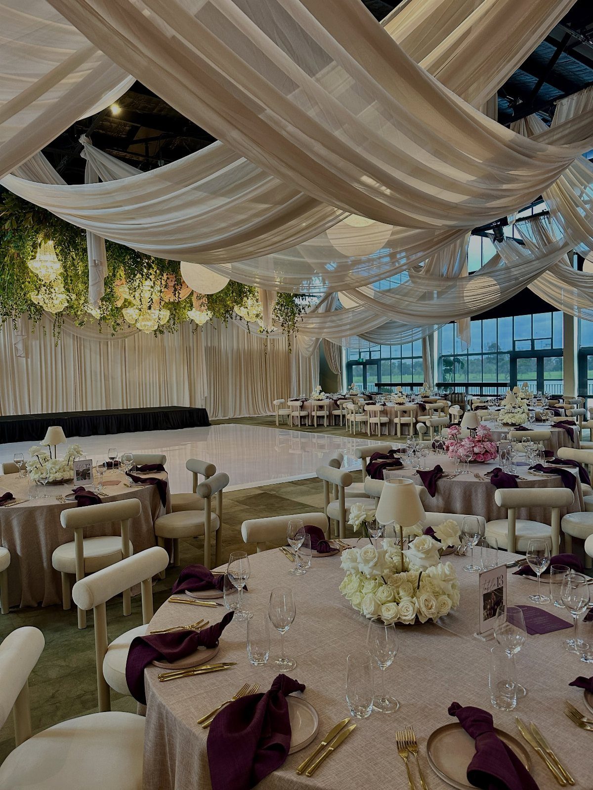 Luxury wedding reception with white ceiling draping, round tables, and glass walls.
