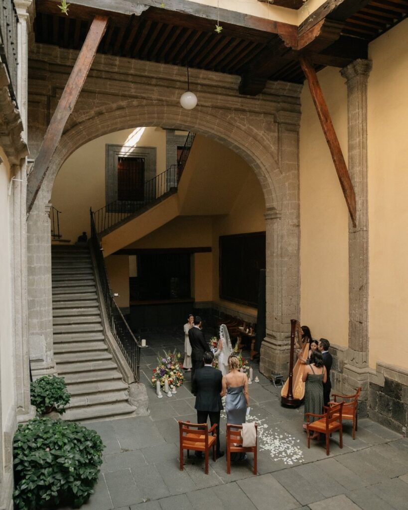 Wedding ceremony in a stone courtyard with a large arch and yellow walls.
