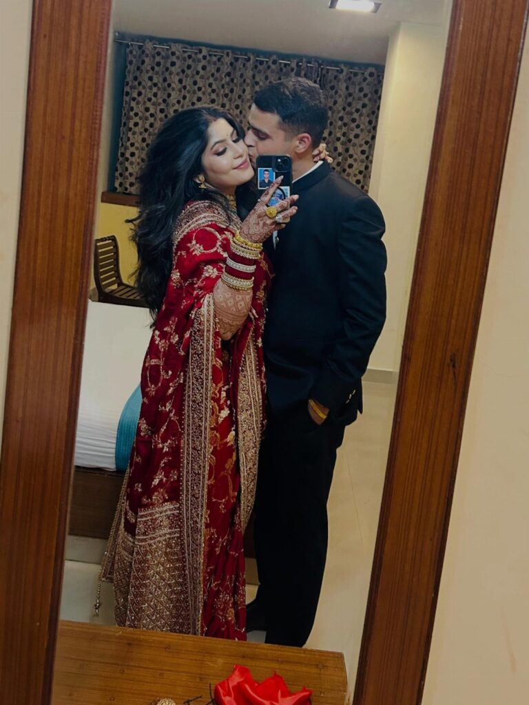 Couple in traditional Indian wedding attire taking a mirror selfie in a room.