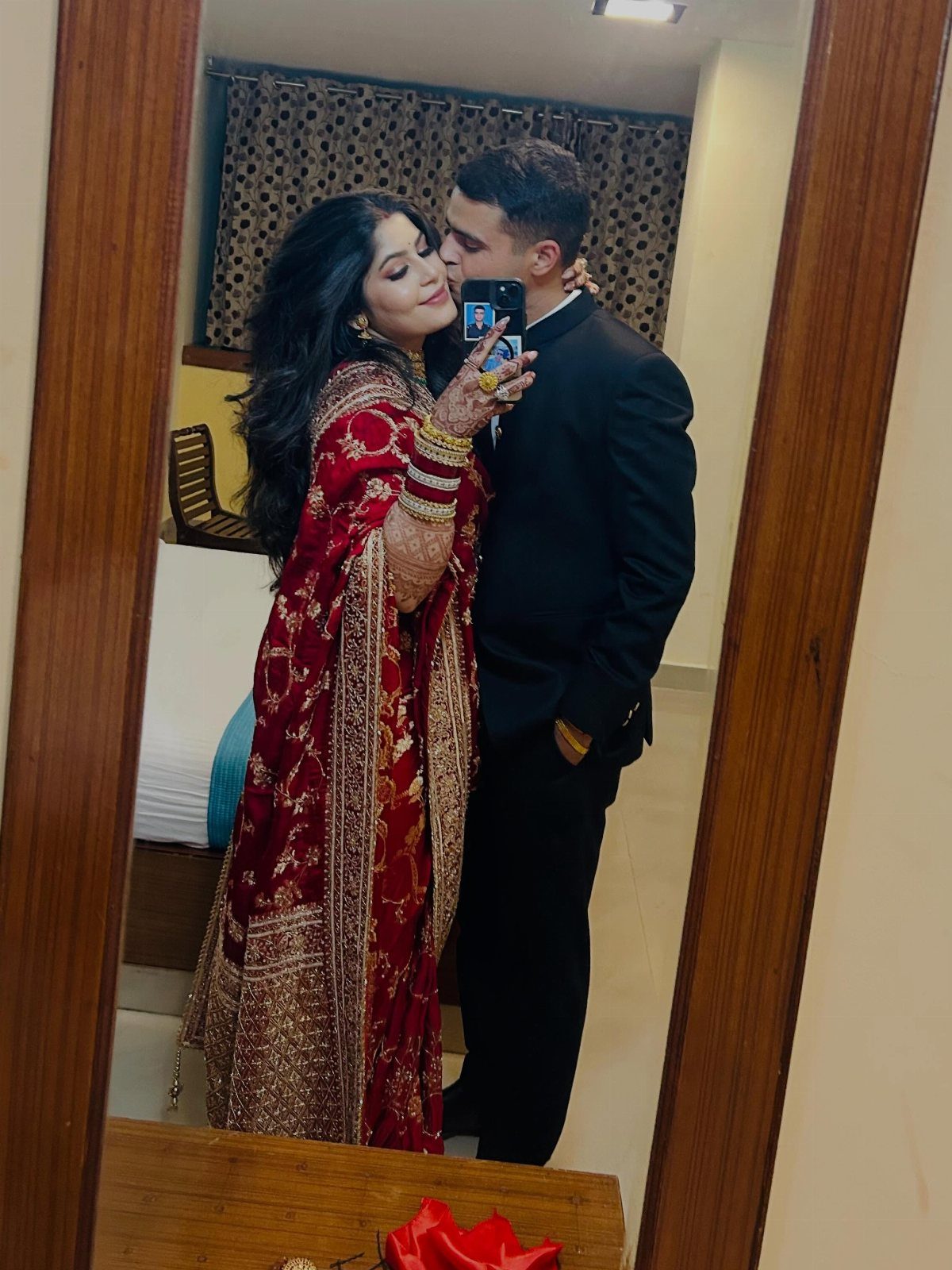 Couple in traditional Indian wedding attire taking a mirror selfie in a room.