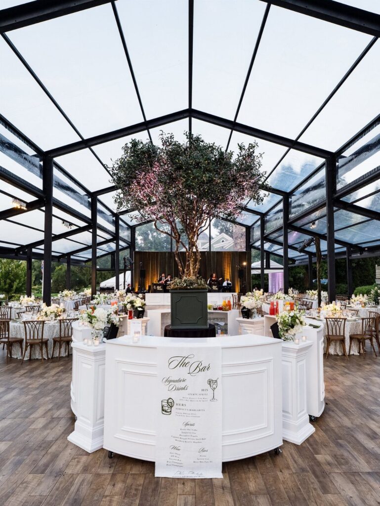 Glass pavilion wedding venue with white tables, floral arrangements, and a central tree.