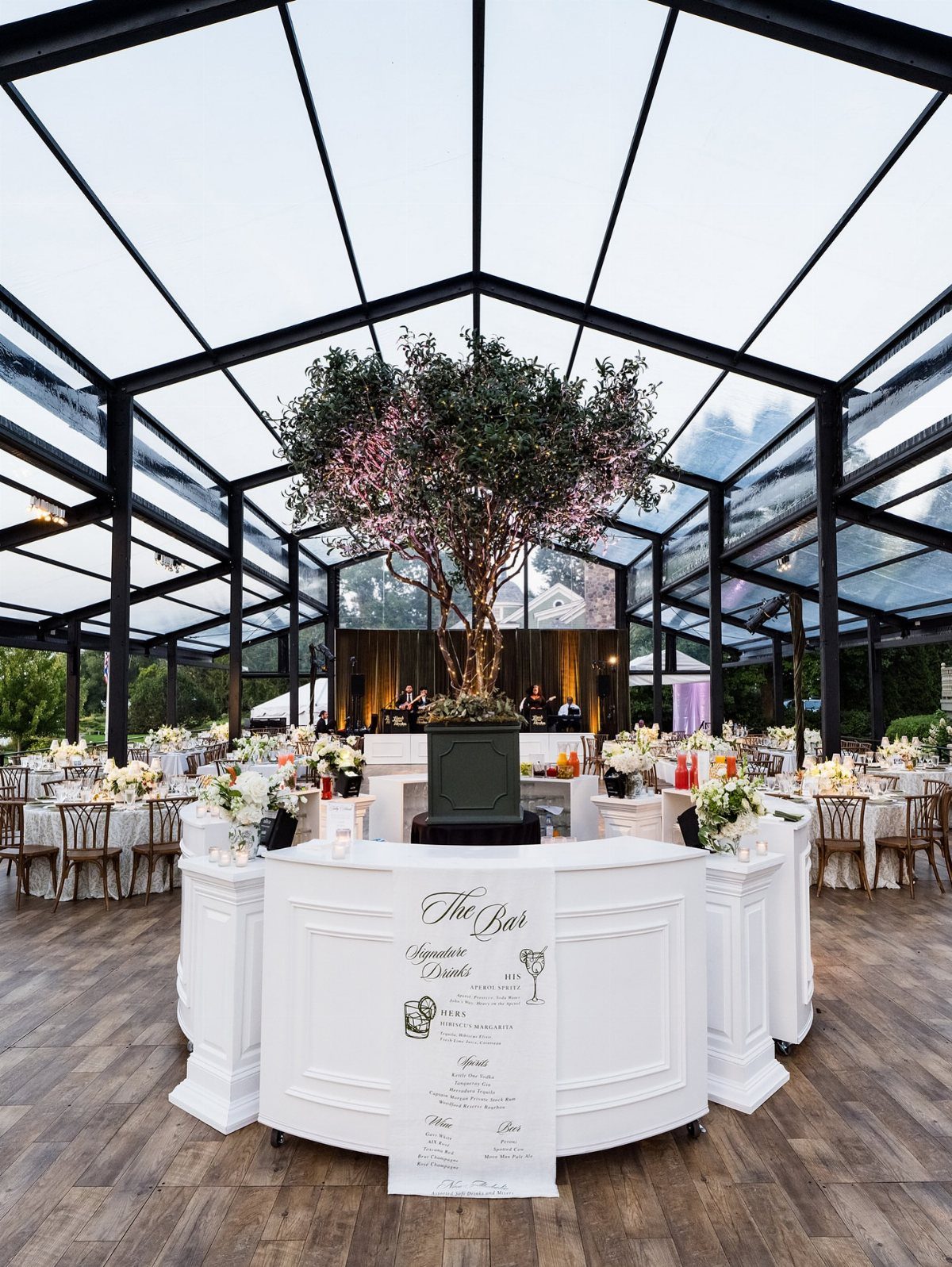 Glass pavilion wedding venue with white tables, floral arrangements, and a central tree.