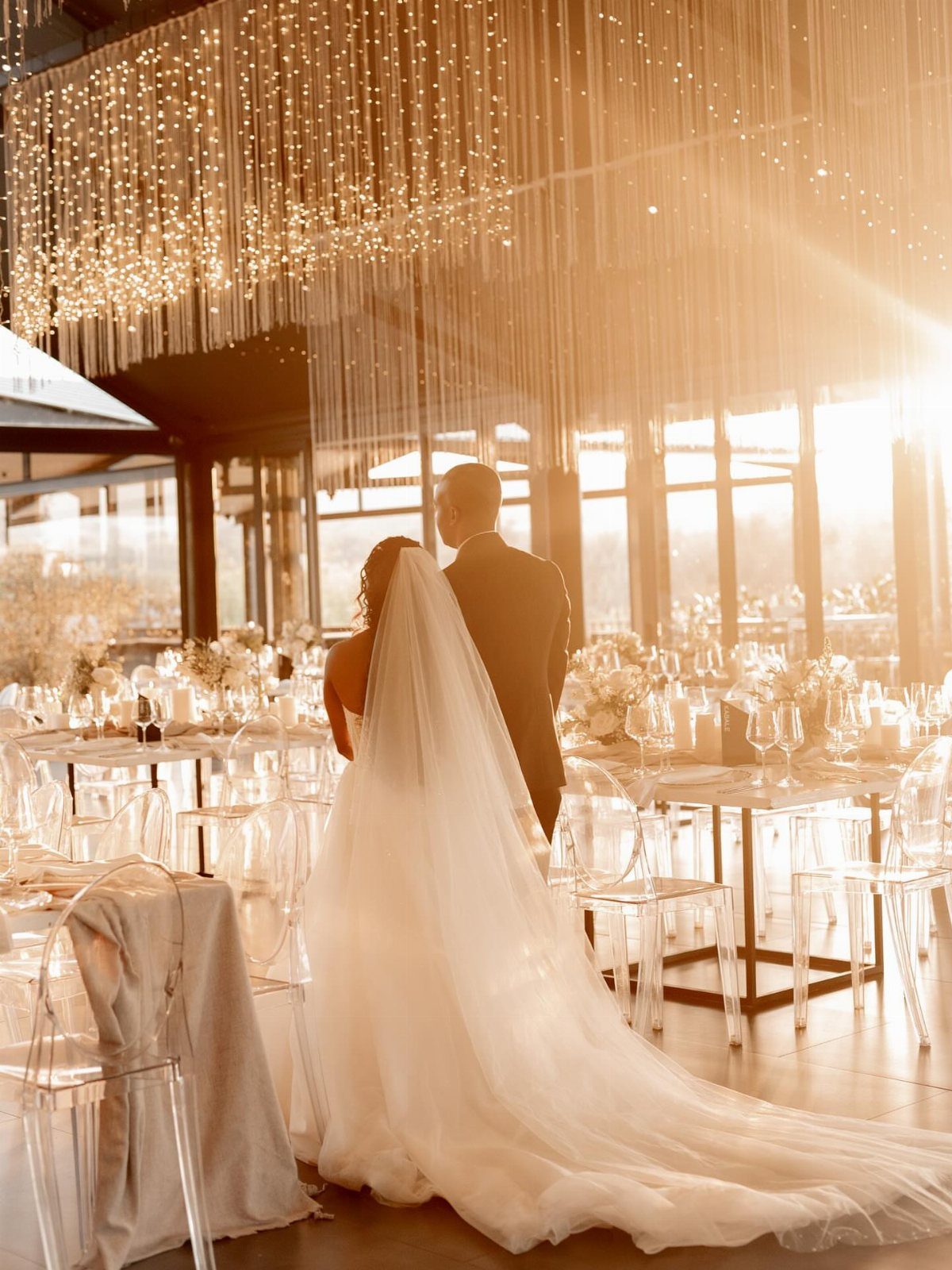 Bride and groom standing in a wedding venue with hanging fairy lights and clear chairs.