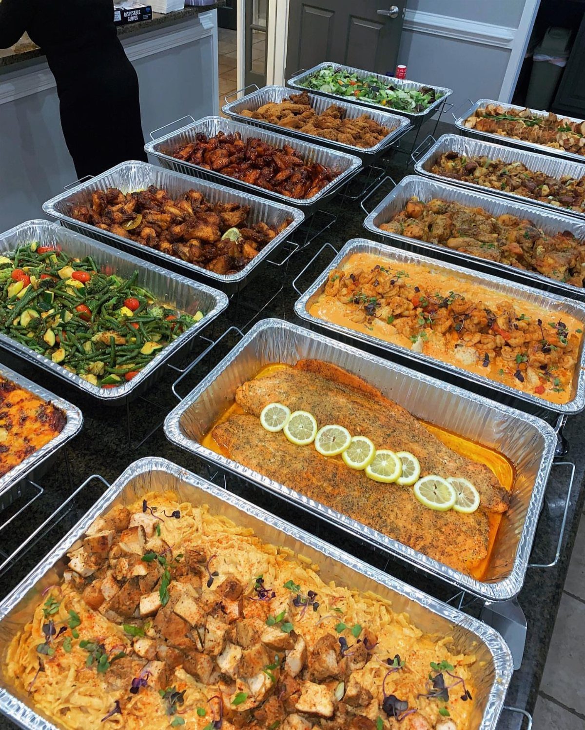Catered food trays featuring salmon, pasta, and vegetables arranged on a dark counter.