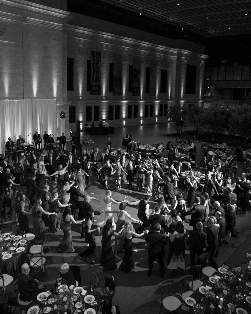 Formal gala reception with guests dancing in a grand stone atrium with high ceilings.