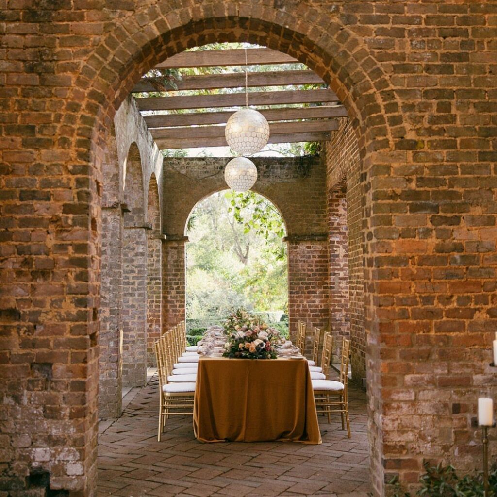 Dining table with gold cloth set within a rustic brick archway courtyard