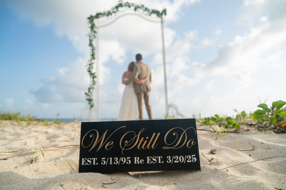 Black sign with gold text on a beach with a couple under an arch.