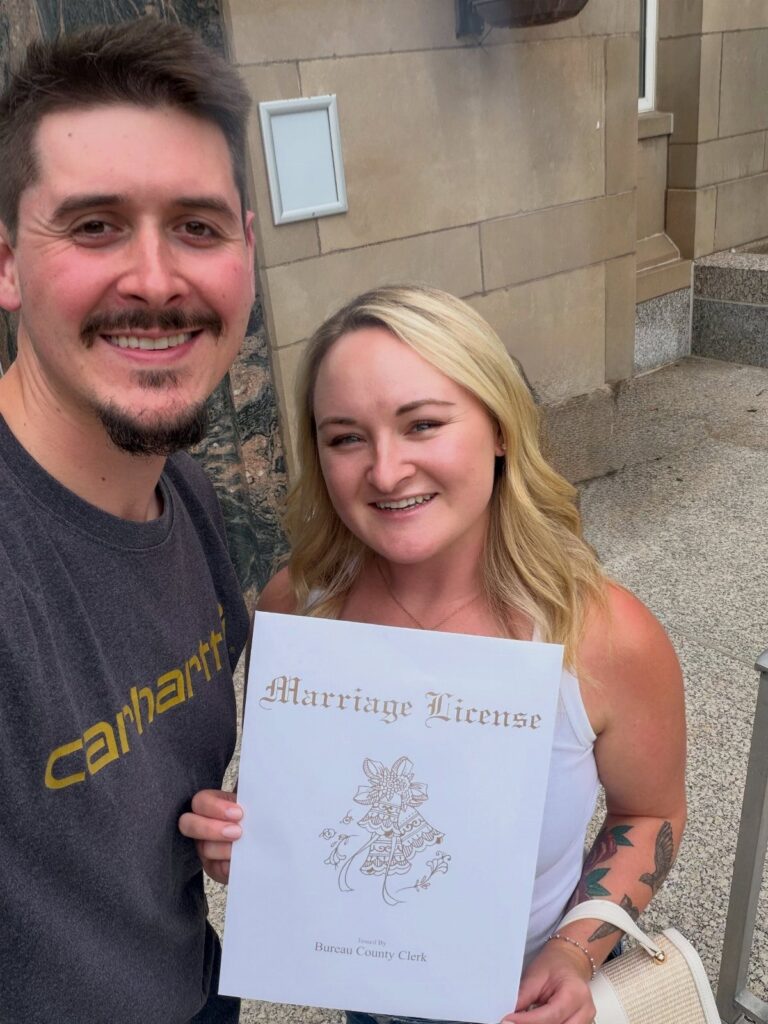 Smiling couple holding a marriage license document in front of a stone building.