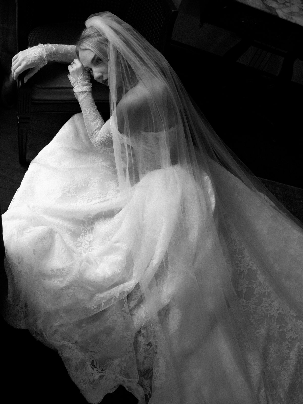 Bride in a lace wedding gown and veil sitting against a dark chair.