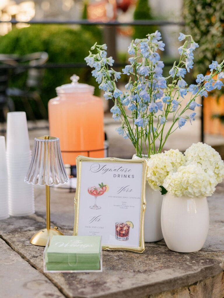 Signature drinks menu and orange beverage dispenser with blue and white flowers outdoors.