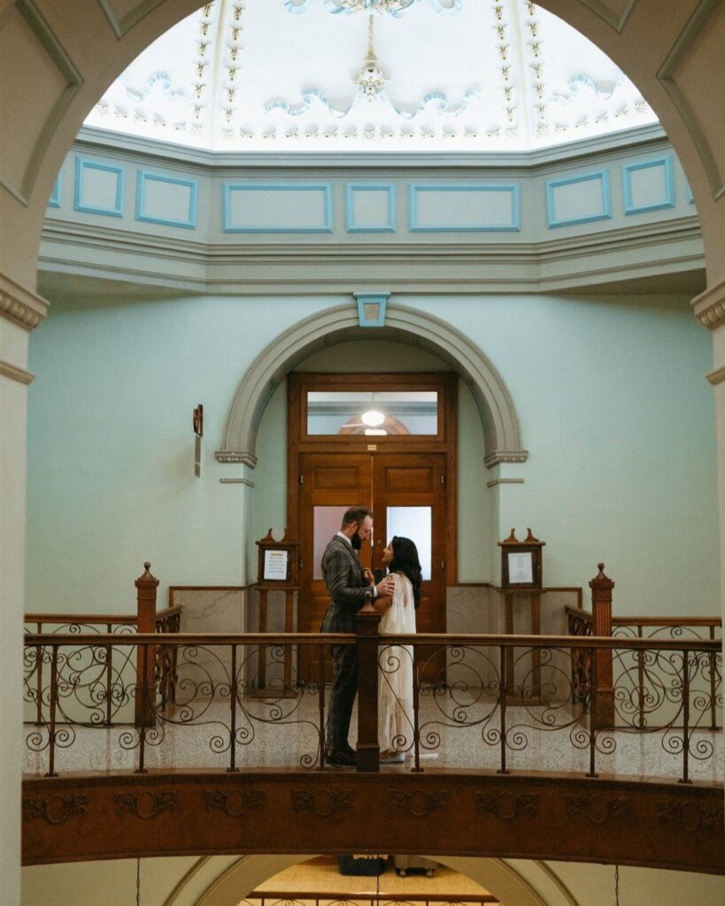 Wedding couple embracing on a balcony inside a courthouse with a domed ceiling.