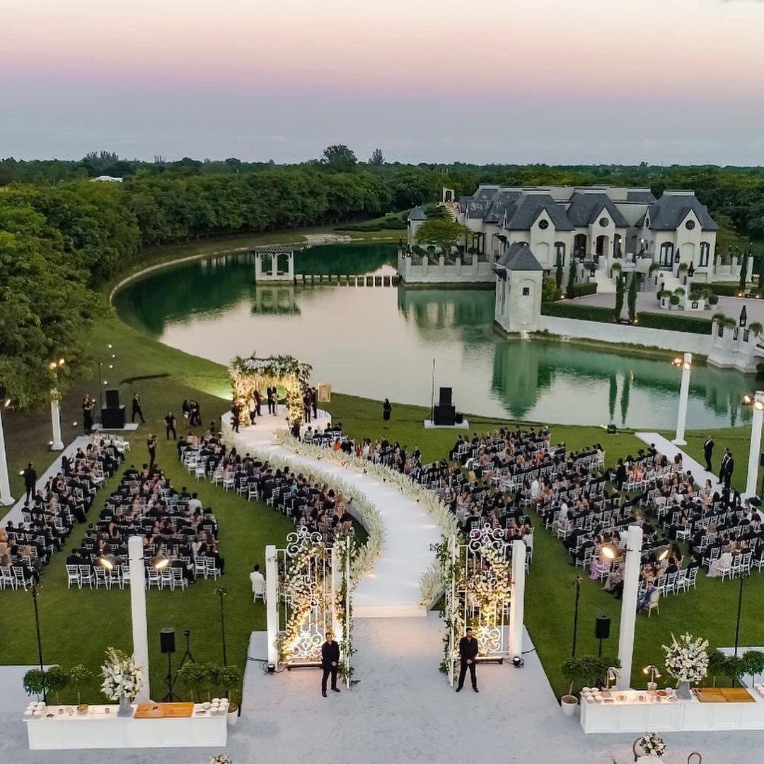 Outdoor wedding ceremony with a white aisle, floral arch, and large estate background.